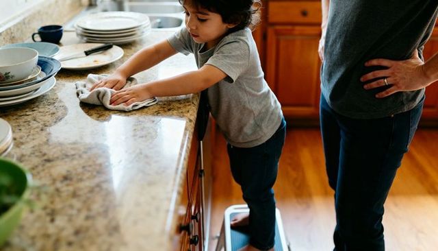 Young child wiping granite countertop while parent supervising on step stool in kitchen