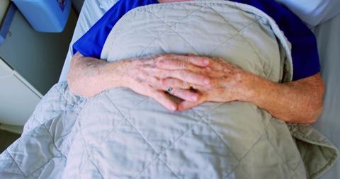 Senior woman resting hands on grey quilted blanket in care facility bed showing comfort