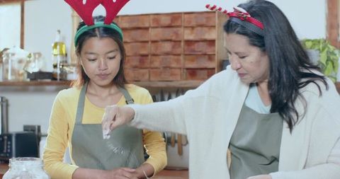 Two women wearing antler headbands baking cookies, sprinkling flour in rustic kitchen