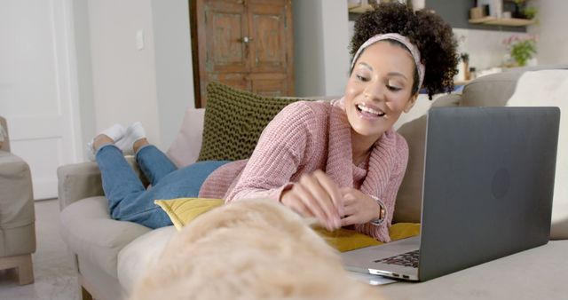 Happy woman lying on a couch while using a laptop and playing with her dog. Perfect for themes on remote work, home comfort, pet ownership, and casual living spaces. Great for blog articles, social media posts, and advertisements related to technology, pet care, and modern lifestyles.