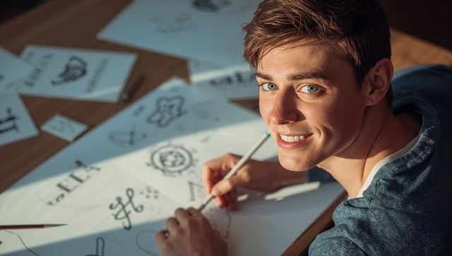 Smiling young designer sketching logo concepts at wooden desk in warm sunlight