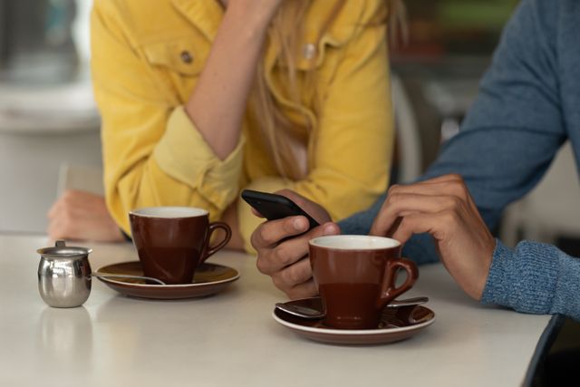 Mid section of a Caucasian couple sitting by a table at a coffee shop, having coffee, enjoying time together and using a smartphone.