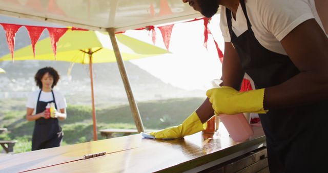 This image shows an outdoor cafe worker cleaning a table while wearing yellow gloves. The sun is shining brightly and a second worker stands blurred in the background holding a beverage. Ideal for use in content about hygiene, sanitation practices in cafes, outdoor dining, and health and safety compliance.