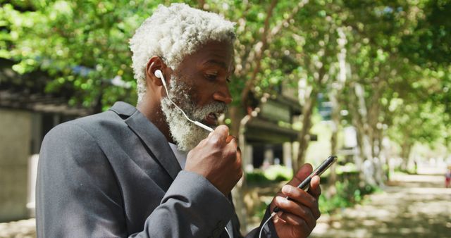 Elderly man with grey hair enjoying his day by listening to music on his smartphone while walking in a park. He is using earphones and dressed in business attire. The greenery and sunlight suggest it is a pleasant summer day. Useful for themes such as technology for seniors, outdoor lifestyles, relaxation, and the modern life of the elderly.