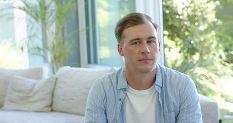 Calm young man sitting on sofa in bright modern living room facing camera through window
