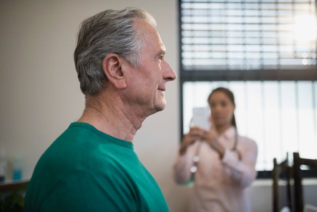 Senior male patient standing in hospital ward while female therapist photographs him. Useful for healthcare, medical treatment, elderly care, and professional medical services themes.