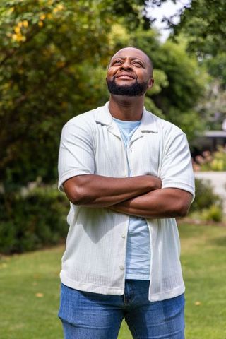 Confident man in 30s standing with arms crossed looking upward in sunny backyard garden