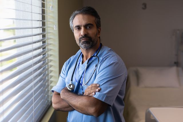Male doctor in scrubs standing with arms crossed in a hospital room, looking confident and professional. Ideal for use in healthcare-related content, medical websites, hospital brochures, and professional profiles.