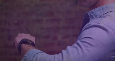 Man checking smartwatch on right wrist wearing light-blue shirt against purple brick wall