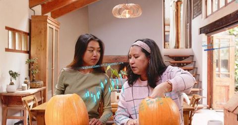 Two women carving pumpkins in sunlit rustic loft with digital finance chart overlay