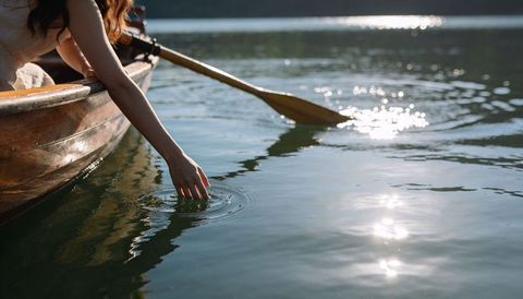 Sitting woman touching calm lake water from wooden rowboat with oar and sunlit reflections