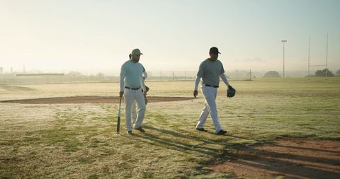 Baseball players walking across dew-covered field at dawn carrying bat and glove