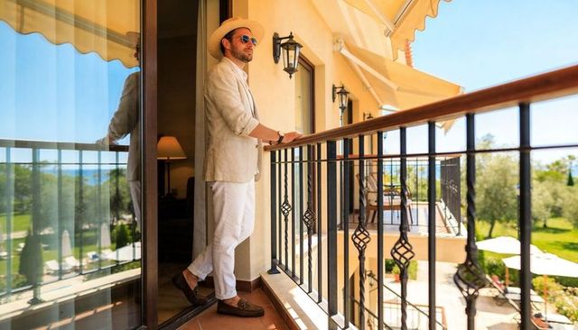 Stylish man leaning on hotel balcony railing wearing light linen blazer and straw hat