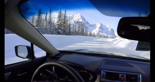 Car interior view of journey through scenic snowy mountain road. Pine trees, mountains, and clear blue sky visible through windshield. Great for themes of winter travel, adventure, road trips, and natural beauty.