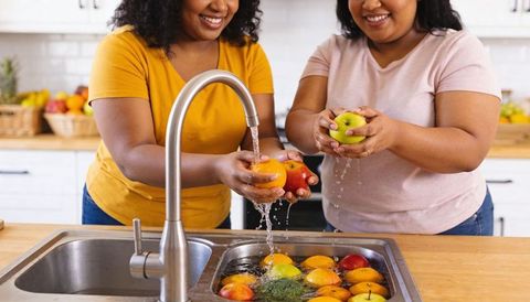 Two women rinsing apples and oranges at kitchen sink smiling during healthy food prep