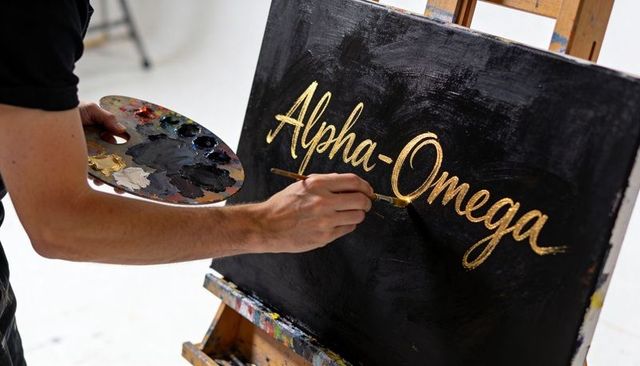 Artist painting gold calligraphy alpha-omega on black canvas, metallic lettering with brush and pale