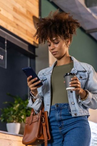 African american woman checking phone holding coffee at urban cafe wearing denim jacket