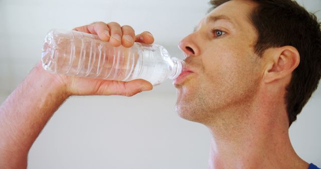 Caucasian man standing and drinking water from plastic bottle in bright gym. Hobby, sport, health and free time, unaltered.
