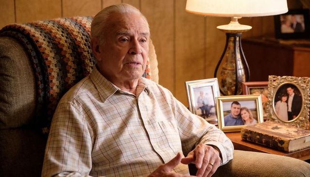 Senior man sitting in cozy wood-paneled living room gesturing surrounded by family photos