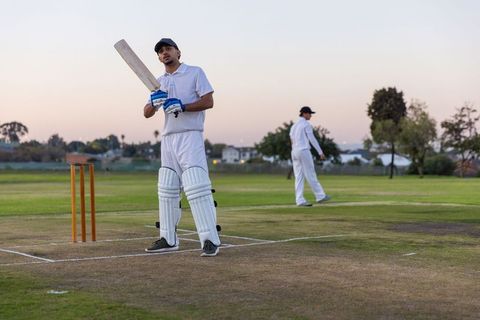 Cricket batsman preparing to bat at dusk on grassy pitch with teammate walking behind