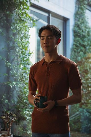 Serene asian man standing in lush courtyard holding coffee mug, morning calm ritual