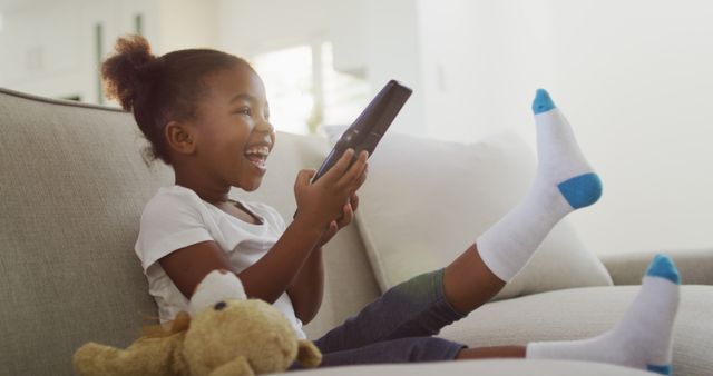 Young girl sits on a couch using a tablet, showing excitement and joy. Great for use in advertisements or articles promoting technology for children, family-focused products, or upbeat home environments. Perfect for educational materials, parenting blogs, or social media campaigns celebrating childhood happiness.