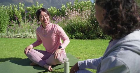Smiling asian woman and partner chatting on yoga mat in garden for outdoor wellness
