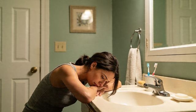 Woman leaning on bathroom vanity resting head on forearms in quiet contemplation