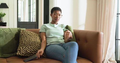 African woman sitting on sofa holding remote, gesturing, relaxing in sunlit living room