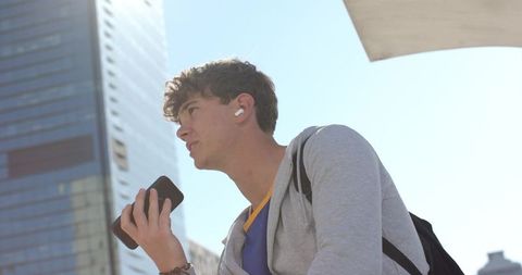 Young man speaking into smartphone while wearing earbuds against modern urban skyline