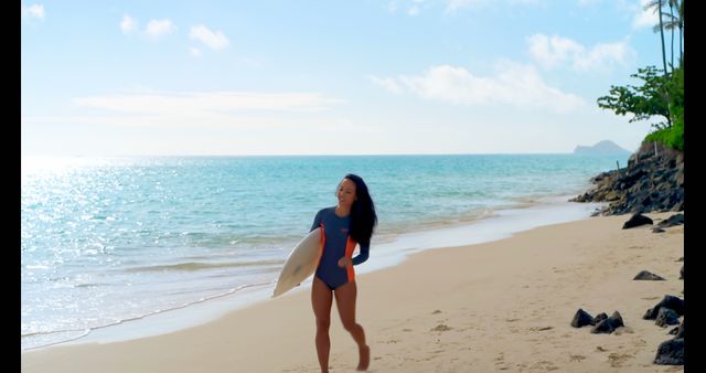 A woman in a navy swimsuit is carrying a surfboard while walking along a sunny tropical beach with clear blue waters and white sand. There are a few rocks scattered in the sand and some trees in the background. This image is ideal for promoting beach holidays, surfing destinations, or summer apparel.