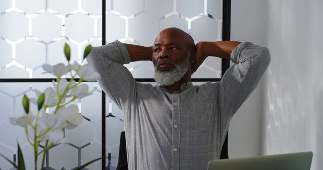 Elderly man in professional setting, taking a break from work by sitting back with arms behind head. Ideal for use in articles about retirement, work-life balance, relaxation techniques, senior professionals, and mental health at work.