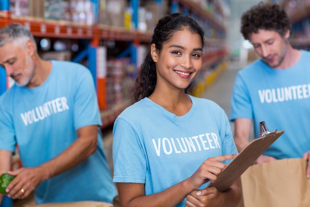 Young woman volunteering in a warehouse, holding a clipboard and smiling. Two other volunteers are seen in the background packing items. Ideal for use in articles or advertisements about community service, charity events, volunteer work, and social responsibility initiatives.