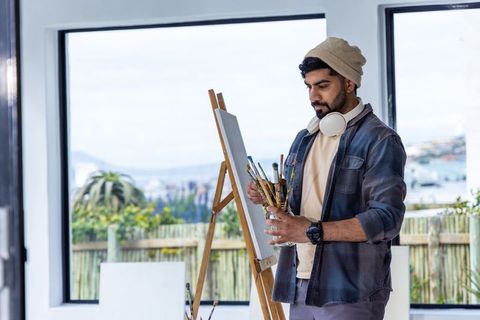 Artist arranging paintbrushes at easel in bright studio with large windows and garden view
