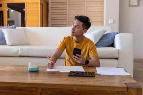 Asian man working at home holding phone and writing on documents on coffee table
