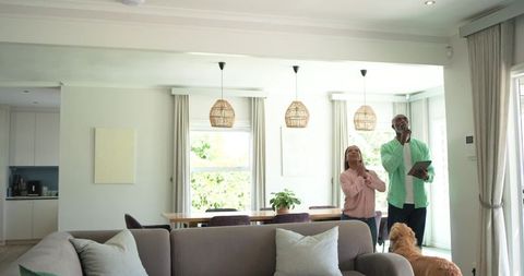 Couple checking ceiling lights while holding tablet in sunlit open-plan living room