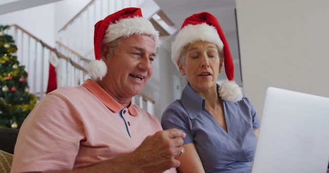 Senior couple sitting in living room wearing Santa hats and video calling relatives on laptop. Suitable for use in campaigns focusing on Christmas holidays, senior technology use, and family communication. Ideal for holiday greeting cards, website visuals, and advertisements.