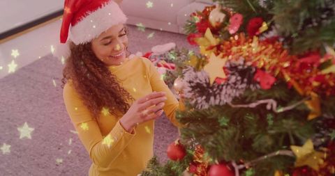 Curly-haired woman in santa hat kneeling and decorating christmas tree with tinsel