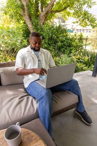 African man working remotely on laptop on lush garden patio lounge with coffee mug