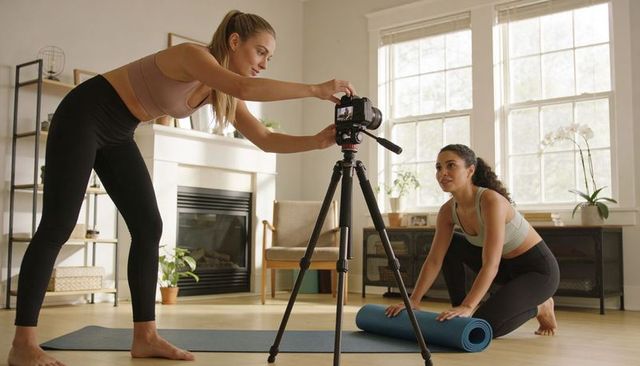 Filming home workout with instructor adjusting camera and participant unrolling yoga mat