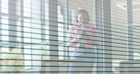 Office professional standing at glass wall checking smartphone through striped blinds