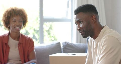 Diverse couple packing moving boxes at home, sitting on sofa calmly and sharing move plans
