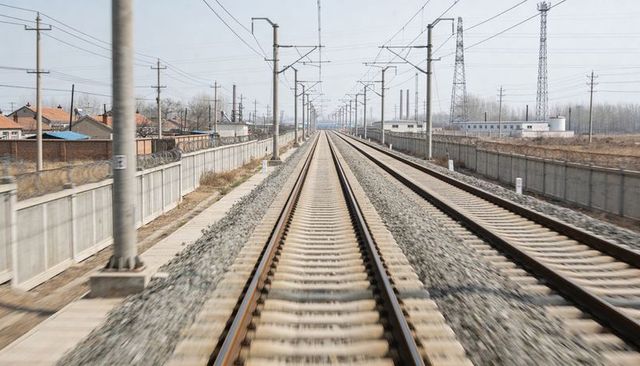 Double railway tracks stretching to horizon in suburban industrial corridor with overhead lines