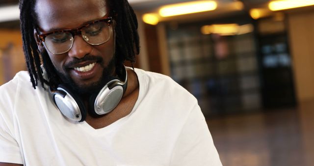 Young man with dreadlocks relaxing indoors, wearing glasses and headphones. Ideal for use in advertisements related to lifestyle, fashion, music, technology, and relaxation. Perfect for conveying themes of enjoyment, style, modernity, and casual living.