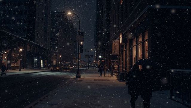 Commuter walking under umbrella on snowy city street at night with warm storefront lights