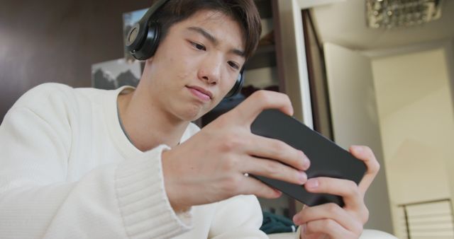 Young man focused on playing mobile game while wearing headphones, ideal for themes related to technology, gaming, entertainment, leisure activities, and youth culture.