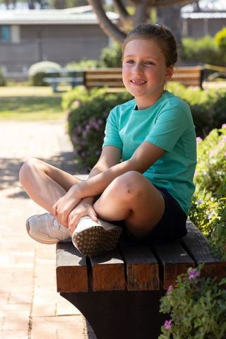 Smiling girl sitting cross-legged on sunlit park bench wearing teal t-shirt and sneakers