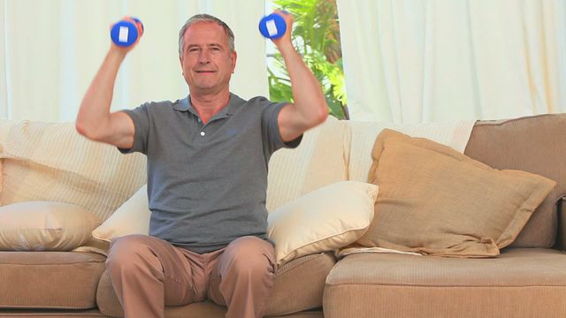 Middle-aged man lifting dumbbells while sitting on couch in living room. Useful for themes related to fitness, healthy lifestyle, home workouts, and exercises for mature adults. Great for websites or materials emphasizing indoor exercise routines and home fitness tips.