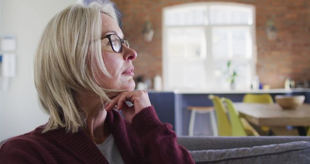 Senior woman with glasses sitting on sofa in cozy living room, looking thoughtfully out window. Perfect for themes related to aging, contemplation, relaxation, home life, wellness, and casual lifestyle.