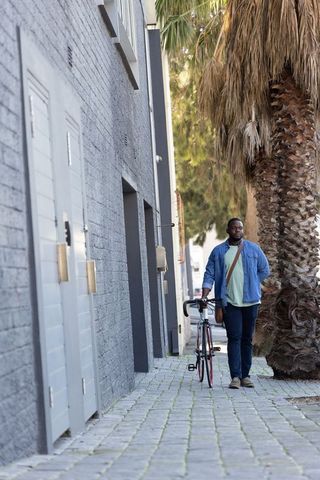 African American man walking with bicycle along palm-lined urban walkway in denim jacket